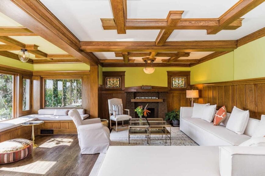 Yellow living room accented with stylish wood beams on the ceiling complementing the half wood panel wall along with a hardwood flooring. It has a huge white- L-shaped sofa that matches the armchairs and the L-shaped window nook seat.