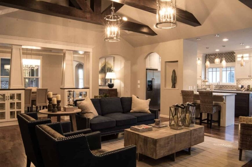 Craftsman living room features a rustic wood coffee table paired with a black sofa set. It is illuminated by cylindrical chandeliers that hung from a vaulted ceiling with exposed wood beams.