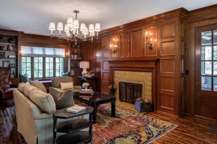 Cozy living room lighted by a lovely chandelier and brass candle sconces mounted to the wood wainscoted walls where a fireplace is fixed.