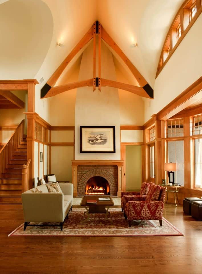 Craftsman living room accented by a triangular wooden beam on a high, vaulted ceiling. It features mismatched sofas over a red vintage rug facing the brick fireplace that warms the room.