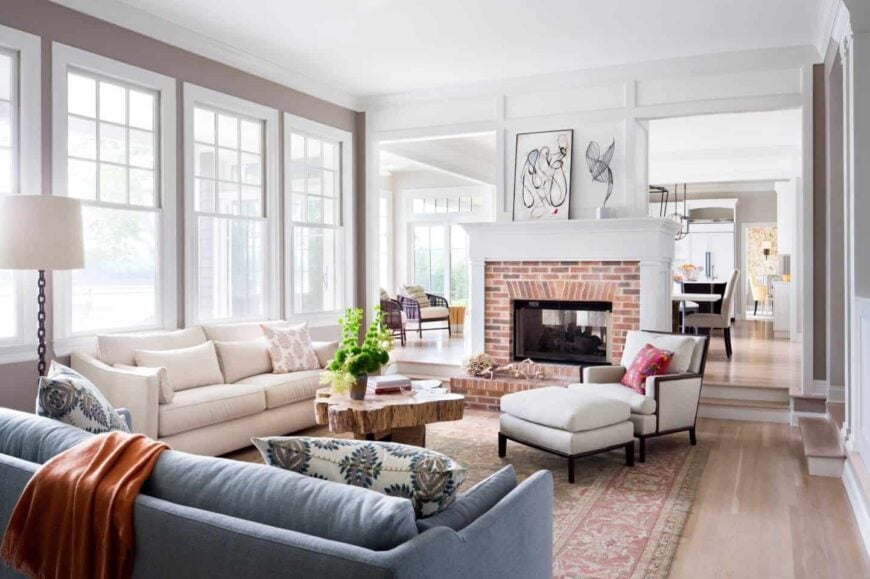 Bright living room with glass windows, white and blue sectionals, brick fireplace and a tree trunk handmade coffee table that sits on a red vintage rug over hardwood flooring.