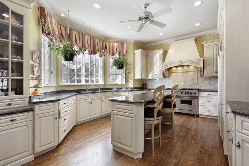 Kitchen features yellow walls with brick tiles backsplash, white cabinets, hardwood floors and a ceiling fan hung over a white island table topped with black marble countertop seating a pair of wooden with black cushion chairs.