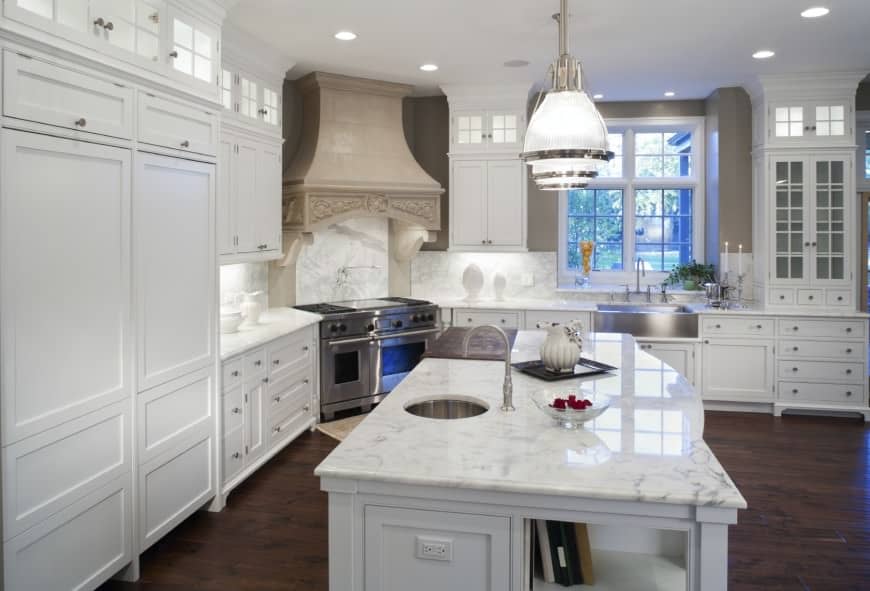 Kitchen with white cabinets, hardwood flooring, big pendant lights, island table with marble countertop and stainless stove with dual ovens and a nice vent hood.
