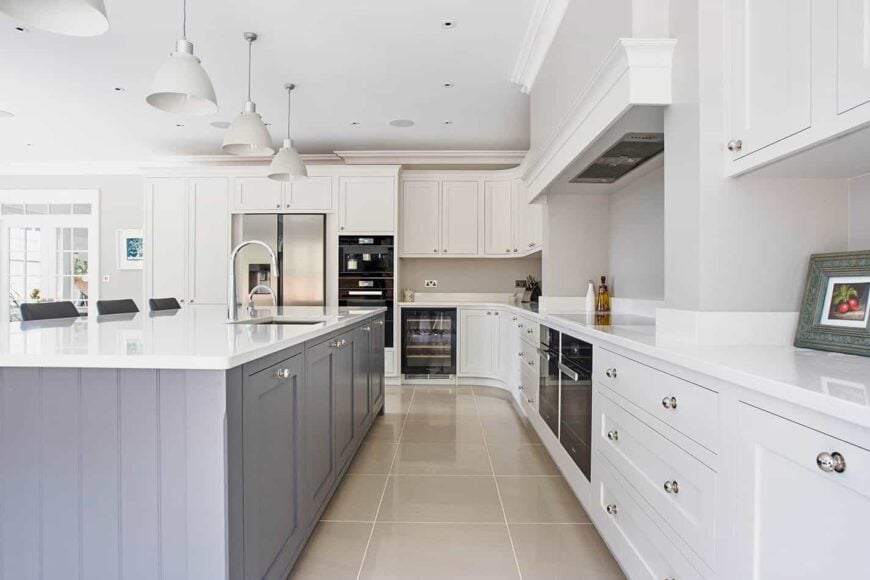 Beautiful cottage kitchen painted with muted gray walls. It features white cabinets, tiled floors and three pendant lights hung over a dusty blue breakfast island topped with white marble.