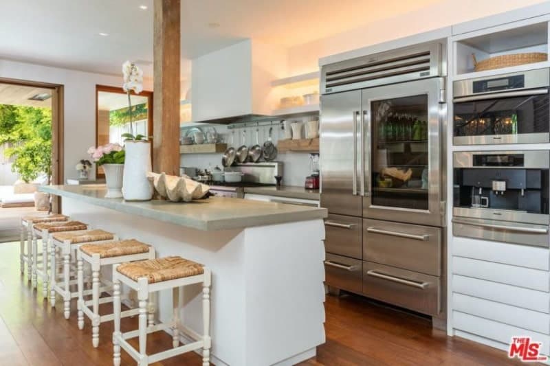 Gorgeous kitchen features a white island table topped with gray marble countertop seating white and brown counter stools, hardwood flooring and stainless steel appliances.