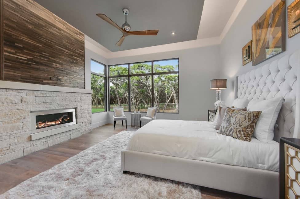 This primary bedroom offers a large luxurious bed set on the hardwood flooring topped by a rug. The room has a fireplace and glass windows.