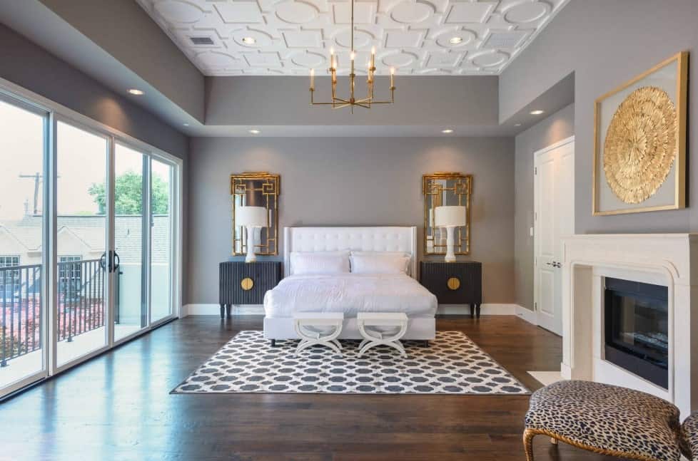 Modern primary bedroom featuring hardwood flooring, gray walls and a fireplace, along with a stunning ceiling lighted by a fabulous chandelier.