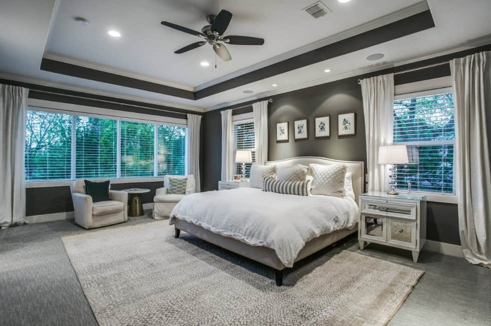 Primary bedroom boasting a white and gray tray ceiling matching the walls and carpet flooring. The room has a large bed lighted by table lamps.