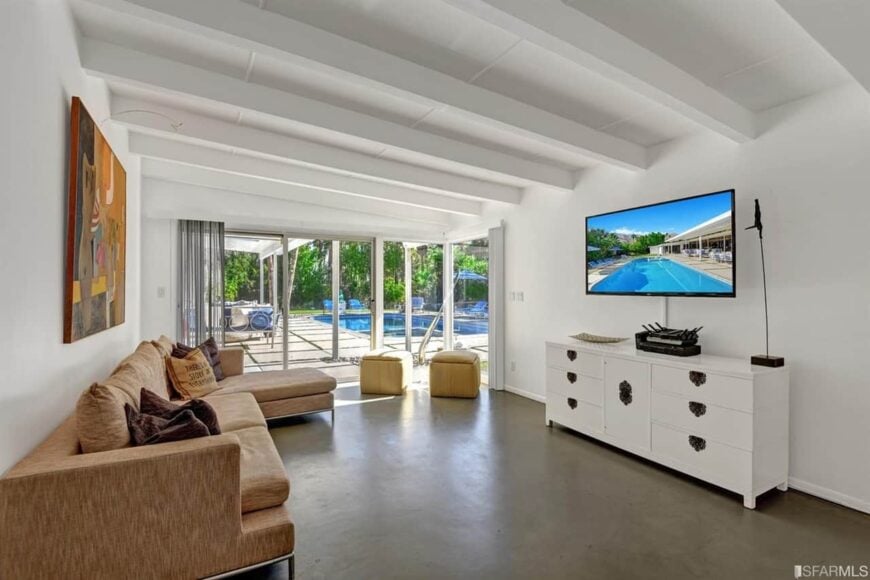 This living room offers a pair of ottomans and an L-shaped sectional facing the white console table and television. It has concrete flooring and glass sliders that open to the patio and the sparkling swimming pool.