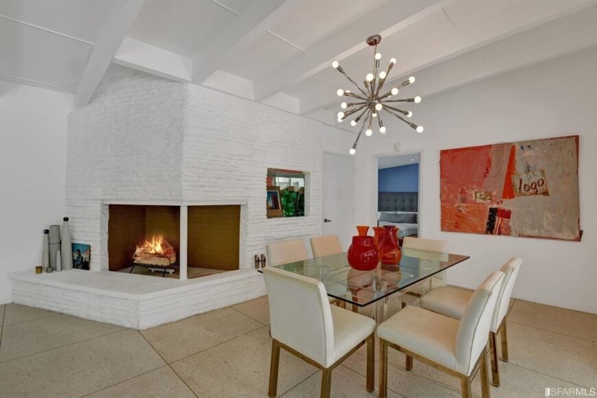 White themed dining room with a striking pendant light, glass table for six, upholstered white chairs, a fireplace, tiled floors.
