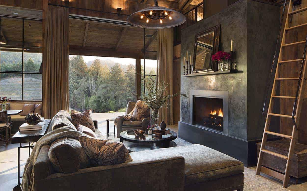 The large L-shaped brown velvet sofa is paired with a matching brown velvet cushioned armchair facing a circular glass-top coffee table by the fireplace that is inlaid within an elegant black column of concrete adorned with a mirror above it.