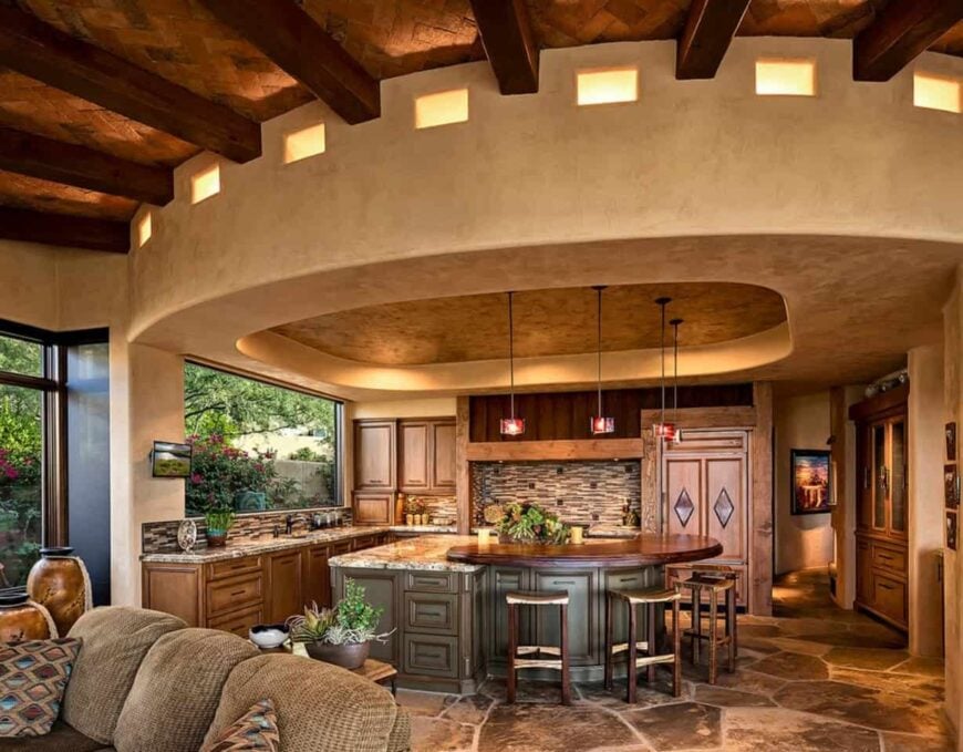 Magnificent kitchen boasts a breakfast island with raised countertop and lighted by pendant lights that hung from a tray ceiling. It has ceramic tile backsplash and beautiful stone flooring.