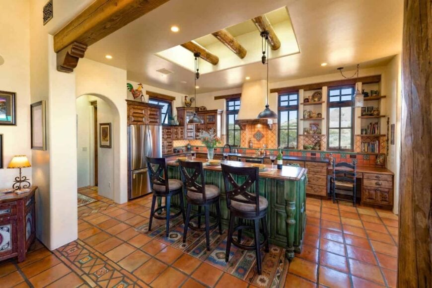 An open kitchen with wooden cabinetry and shelves along with slate flooring. It has a green breakfast island paired with cushioned counter stools.
