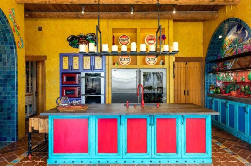 An oversized industrial chandelier hung over a two-toned breakfast island that's fitted with a sink and red fixtures in this colorful southwestern kitchen.