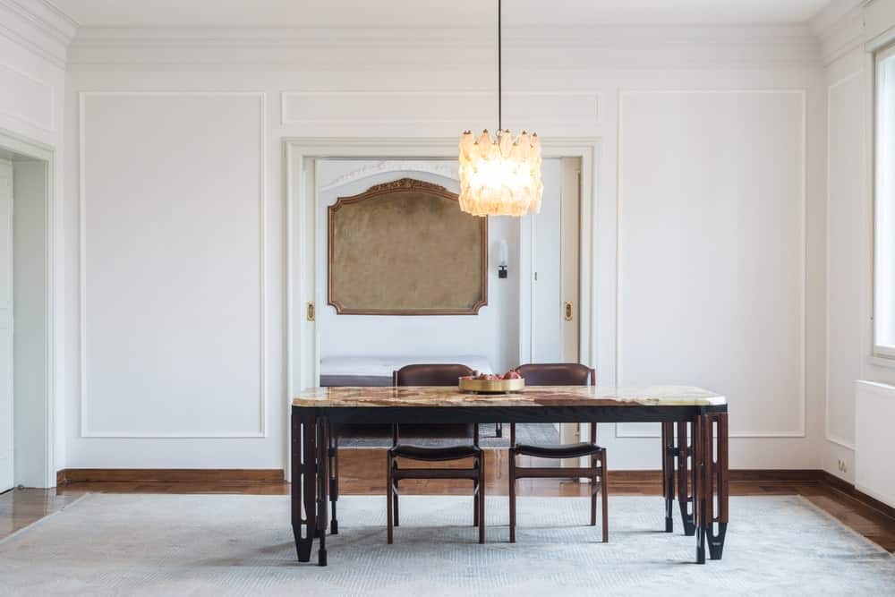 White dining room featuring a large rug covering the smooth brown flooring. The table looks absolutely elegant, perfect together with the ceiling lighting.