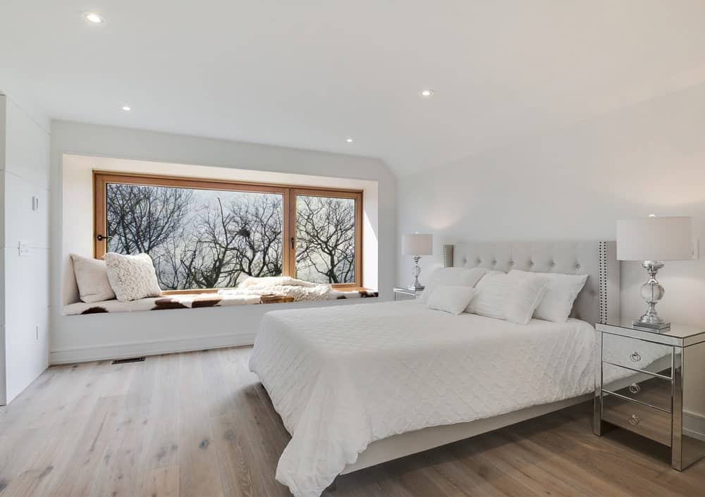 This is a close look at the primary bedroom with a alrge white bed flanked by mirrored bedside drawers with decorative desk lamps. These are complemented by the large window on the far side that has a reading nook.