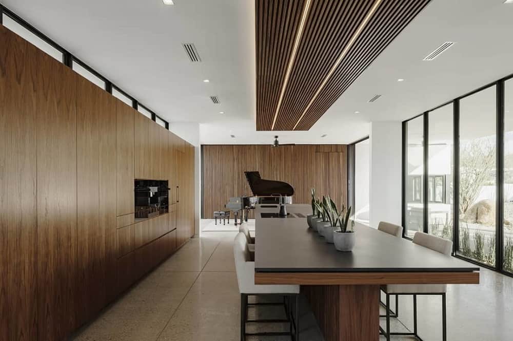 This is a close look at the kitchen that has consistent dark wood tones on its cabinetry and kitchen island that has an attached table topped with a wood-paneled ceiling and black counter.