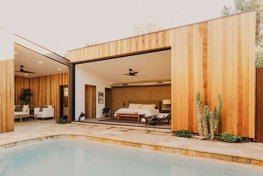 This is a view of the bedroom from the vantage of the pool that has a large glass wall that opens up the interior to the pool area. The bed has a large wooden platform bed adorned with a brick wall behind the headboard.