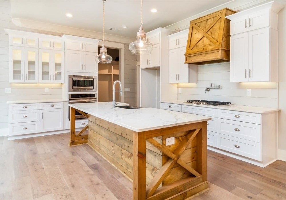 This is a close look at the Farmhouse-style kitchen with a kitche island that blends with the hardwood flooring and the vent hood above the cooking area. These are then complemented by the white cabinetry and white countertops.