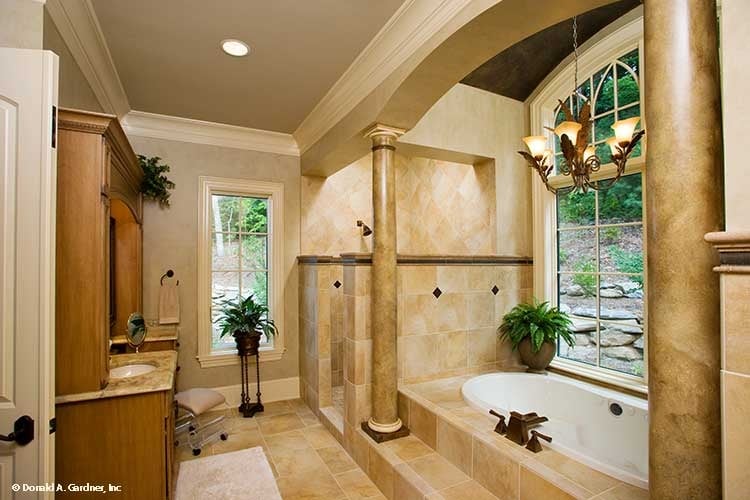 The primary bathroom has a large bathtub placed in a large alcove with an arched window, a chandelier and pillars that pair well with the beige tone of the bathroom.