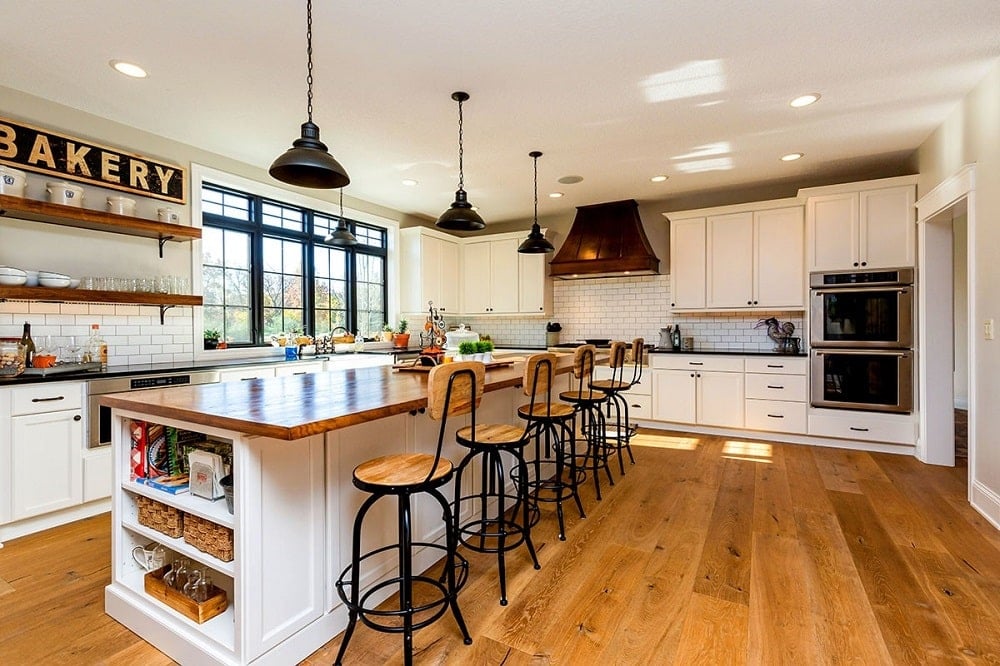 This is the large Farmhouse-style kitchen with a large kitchen island topped with pendant lights and paired with multiple stools that match the hardwood flooring and wooden countertop.