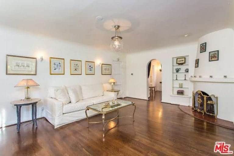 A clean living room designed with wall frames and lighted by a glass globe pendant. It includes a white sofa in the middle of end tables with lamps and a metal coffee table over hardwood flooring.