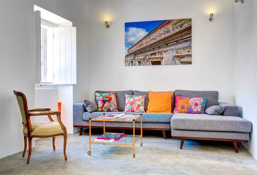 A lovely canvas painting mounted above a gray sectional sofa in this simple living room. It is accompanied by an orange glass top coffee table and upholstered chair over concrete flooring.