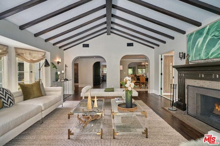 Magnificent living room boasts open arched doors and a cathedral ceiling with exposed wood beams. A pair of glass coffee tables stand in the middle of a white sofa and fireplace.