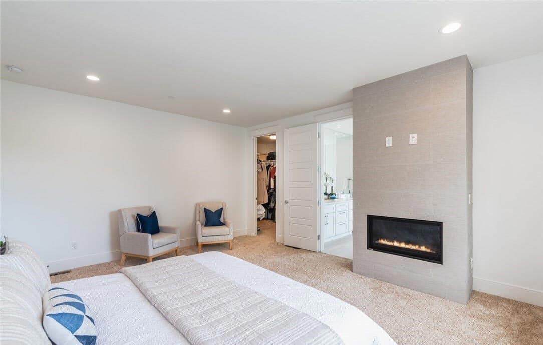 Primary bedroom with a modern fireplace and a sitting area furnished with beige armchairs.