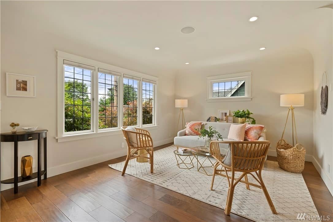 A white patterned area rug tops the hardwood flooring of this Craftsman-Style living room. It is paired with a couple of rustic wooden chairs and a wicker basket beside the grayish sofa that goes beautifully with the white walls and white ceiling. The room is brightened by French windows and a couple of standing lamps flanking the sofa.