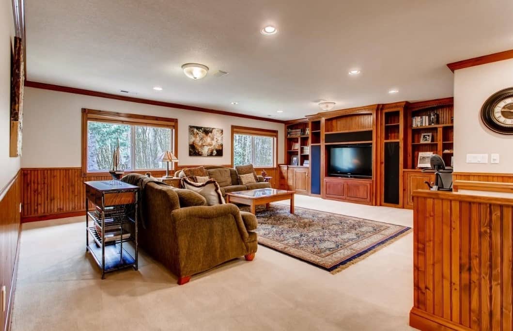 The beige carpeted floor is topped with a colorful patterned rug paired with a pair of inviting velvet sofas. The built-in cabinets that also serve as TV housing and shelves seem to blend into the glass windows and walls with its varnished wooden beauty.
