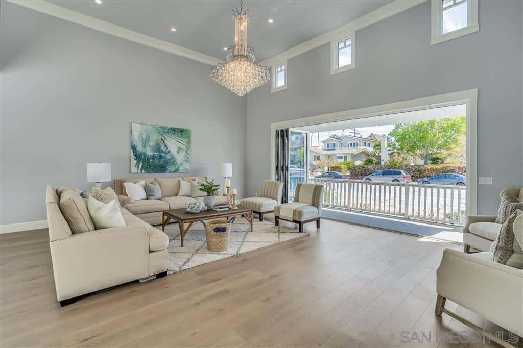The airy quality of this living room is due to its high ceiling and stacking glass doors. This is augmented by the light bluish hue of the walls and a brilliant crystal chandelier hanging over the light grayish sofa chairs matched with a patterned rug.