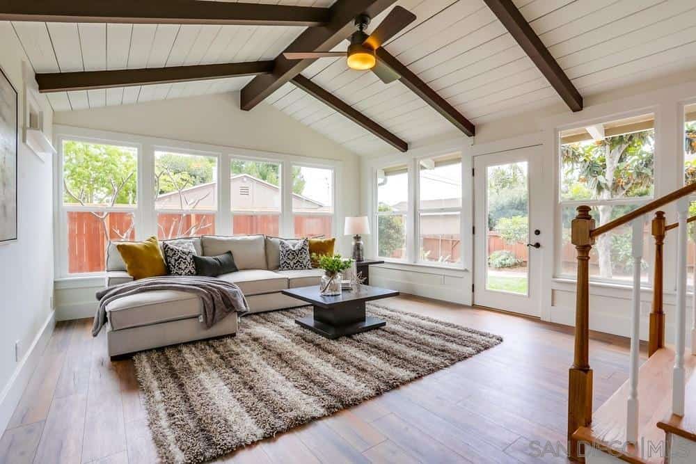 The white cathedral ceiling of this Craftsman-Style living room has wooden exposed beams that contrast the walls that are dominated by glass windows and door. A beautiful striped rug is placed over the wooden floor and paired with a massive L-shaped sofa.