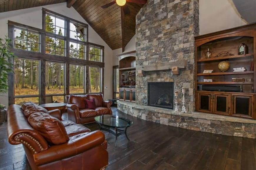 The leather sofas are facing a wall dominated by an extensive stone structure built with built-in wooden shelves flanking a fireplace. The white wall on its side has floor-to-ceiling glass windows that offer a fantastic view of the forest.