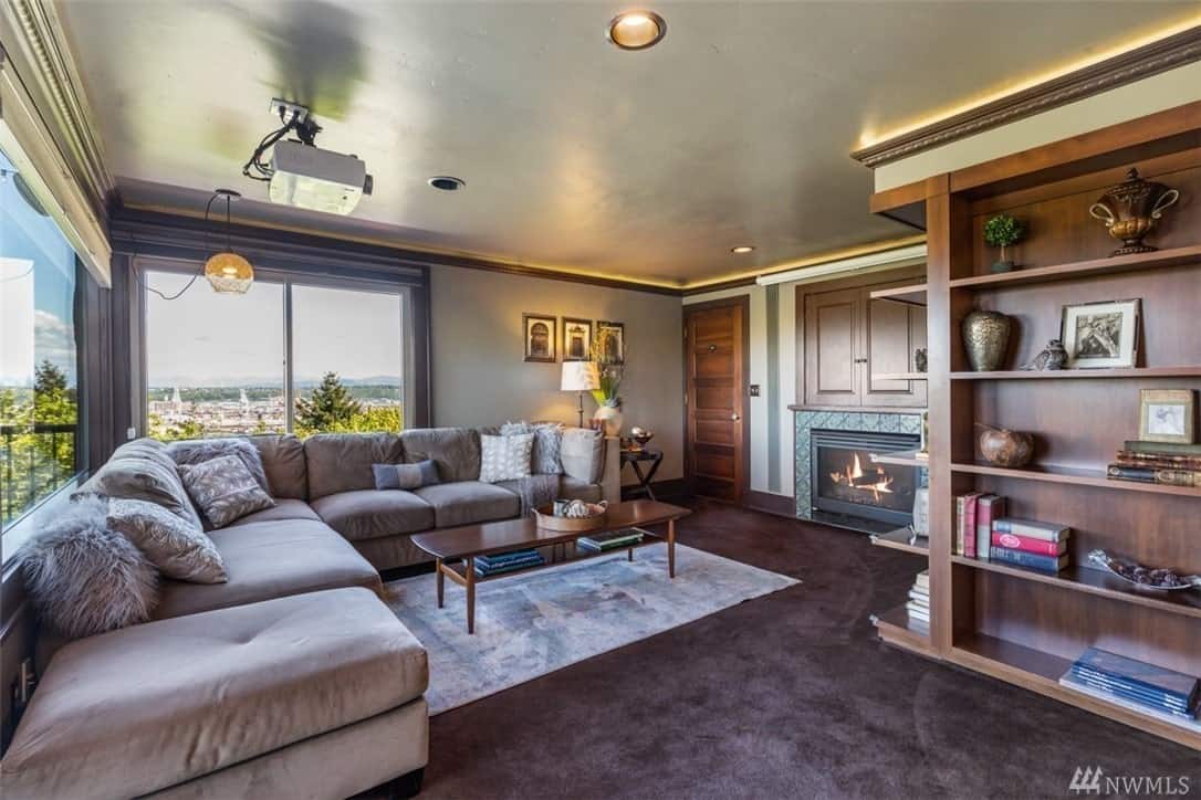This is a traditional Craftsman-Style living room with a modern touch from its ceiling-mounted projector. A massive gray L-shaped sofa faces the fireplace with borders of patterned tiles that contrasts the dark tones of the carpet and wooden TV cover above it.