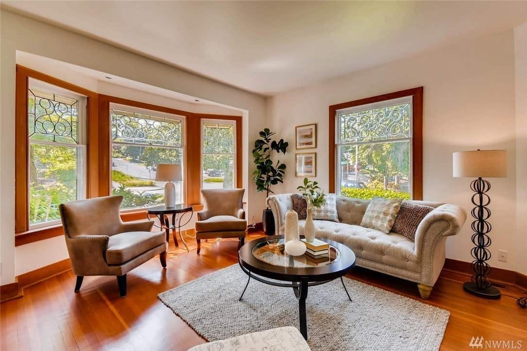 This is an airy Craftsman-style living room with abundant natural lighting coming from its massive glass windows that surround the cushioned sofas. The hardwood flooring is topped with a woven area rug and dark wooden circular coffee table that goes well with the side table and wooden frames of the windows.