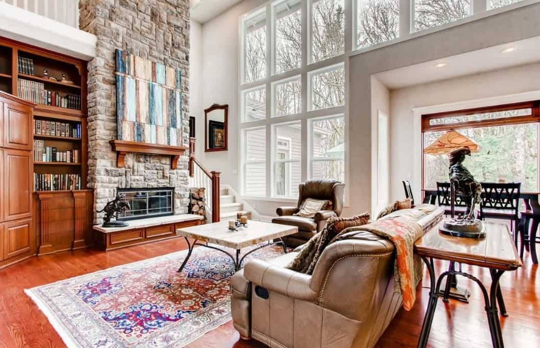 A massive gray stone chimney rises up from the small fireplace that brightened up by the floor-to-ceiling glass windows in this spacious Craftsman-Style living room. Above the fireplace, A colorful abstract painting is mounted which contrasts well with the wood floors, shelves and cabinets. The leather sofas are paired with a rectangular coffee table on a colorful patterned rug.
