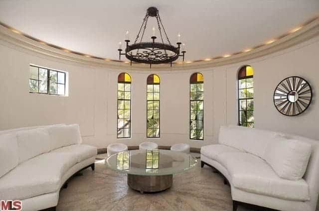 Identical white sofas with a glass top table illuminated by a wrought iron chandelier surround this circular living room.