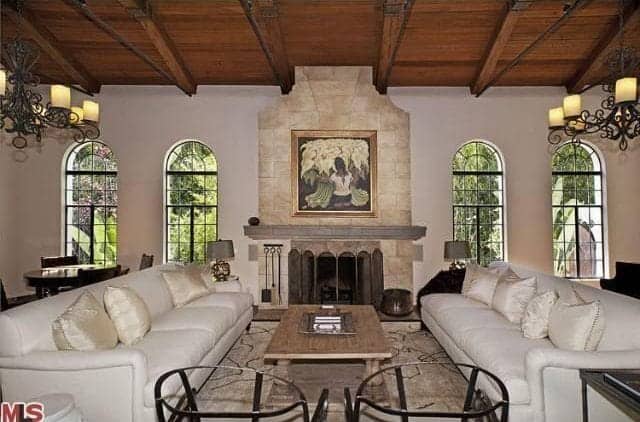 Classic living room with a symmetrical design. It includes white facing sofas and a pair of matching armchairs lighted by vintage chandeliers that hung from a wood beam ceiling.