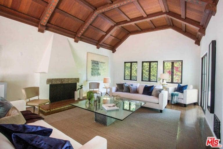 Vaulted ceiling with exposed wood beams covered this white living room. It has matching sofas and chairs with a glass top center table facing the fireplace.