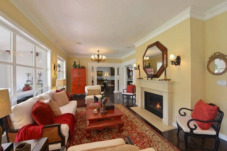 Yellow living room with a pop of red color from the pillows, blanket, rug and redwood coffee table. It has a fireplace underneath a wooden octagon mirror and faces the white sofa.