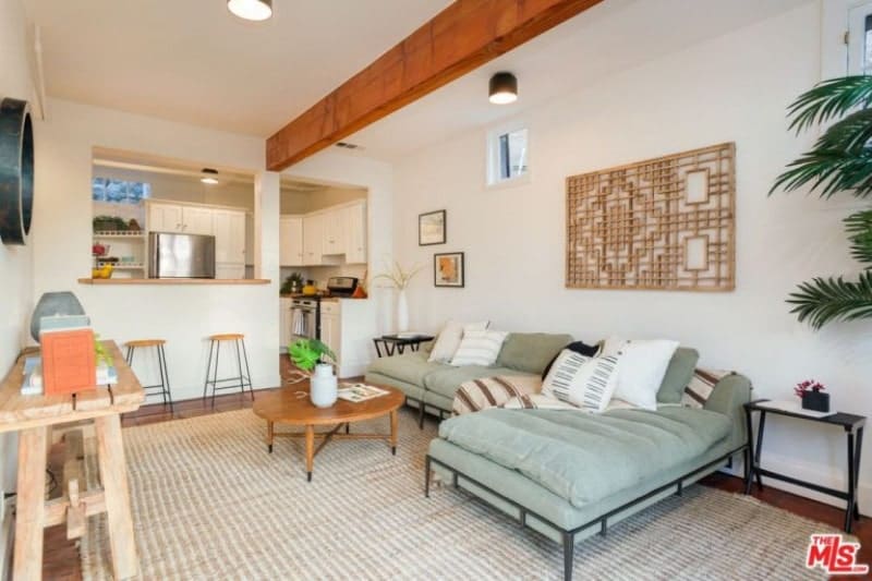 Fresh living room lined with a wood beam from the white ceiling. It is designed with a wood carved wall art mounted above an L-shaped sectional sofa that sits on a striped rug.