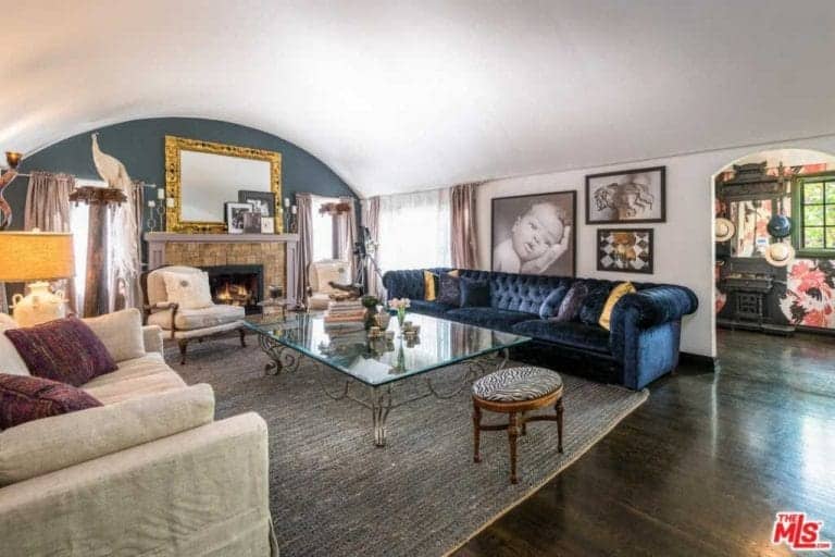 This living room showcases a blue gray arched wall fitted with a brick fireplace topped with gold mirror and frames. It displays a gallery art wall above a deep blue tufted sofa paired by a glass center table and mismatched chairs.