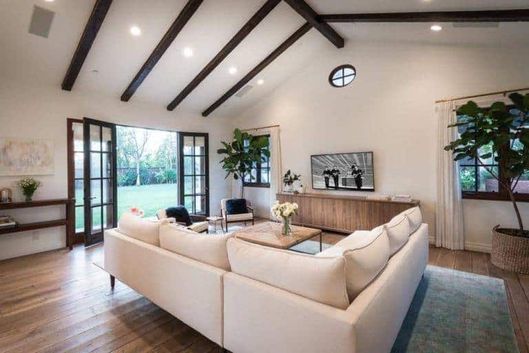 White living room lined with dark wood beams and illuminated by recessed ceiling lights. It includes wood framed windows and doors that bring fresh air and natural light in.