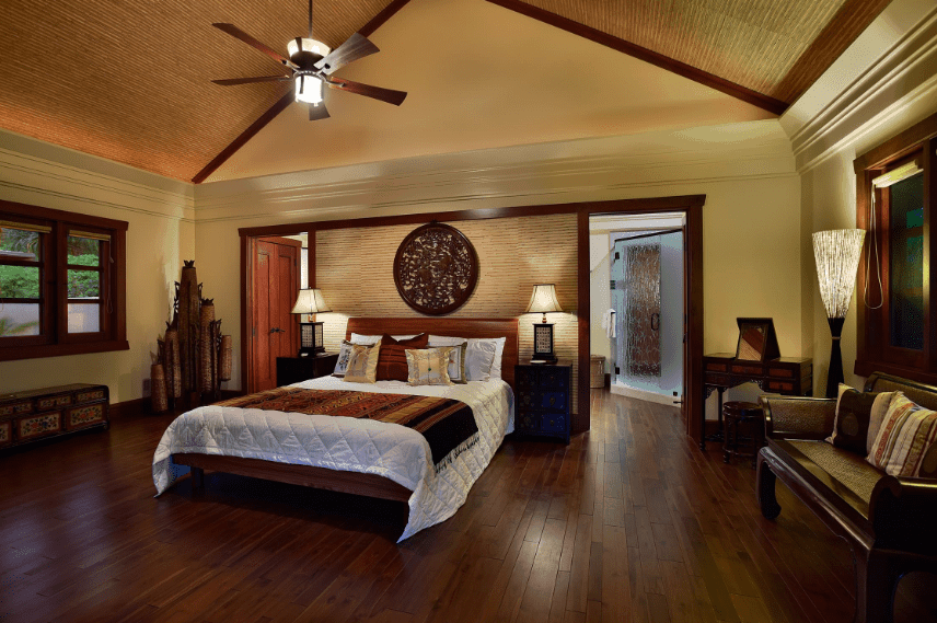 This is a warm and cozy Spanish bedroom that features different hues of wood. The wooden arched ceiling has the same striped pattern as the wall above the headboard where a wall-mounted dark wooden artwork dominates. This dark wooden element is also present in the wooden floors, side tables and even at the wooden sofa by the window.