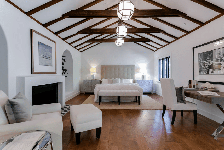 This is a predominantly white Spanish bedroom with white walls, white bed, white chairs, and a white fireplace. This allows the exposed dark wooden beams of the arched ceiling to pop out along with the wooden floor and legs of the pieces of furniture. The whole room is illuminated by three gorgeous hanging polygon lights.