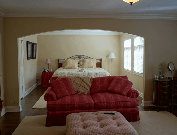 The beige walls and rugs of this Spanish bedroom give it neutrality that is contradicted by the stark color of the sofa by the foot of the bed and the linen covering the side table. The element that balances them is the dark wooden color of the floor, cabinet and wall-mounted frames together with the natural light of the windows.
