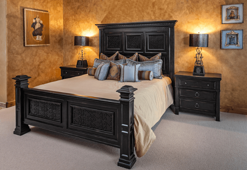A very simple yet dramatic bedroom with a dark wooden bed frame that perfectly pairs with the bedside drawers and the dark lamps on top of them. The cloudy pattern of the wall and the monotone carpet offers a counterbalance to the wall-mounted religious artwork.