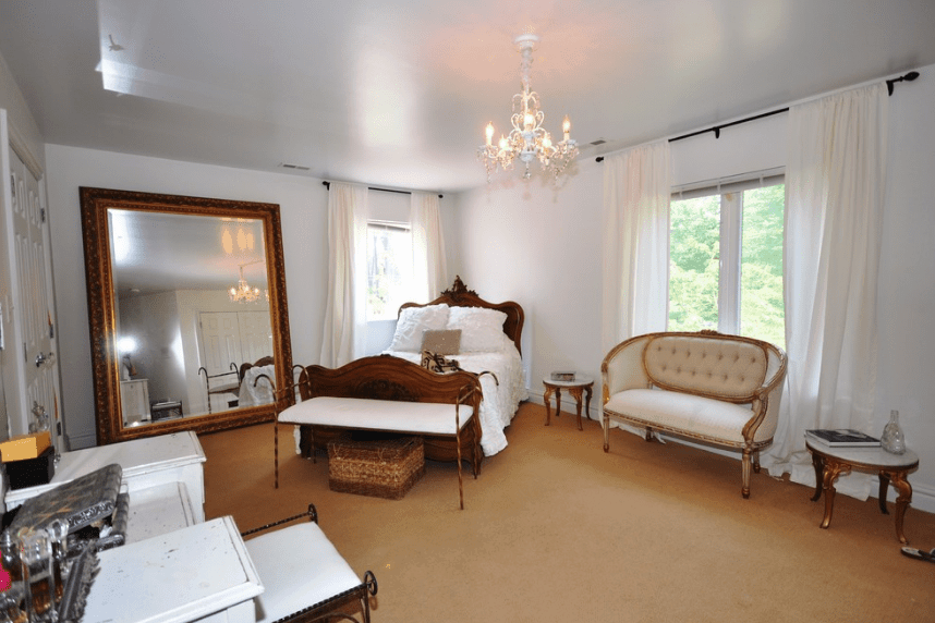 This is a bedroom of grandiose beauty due to its big spacious floor and grand mirror with brass borders that towers over the side of the bed. The white color of the walls and curtains give the room an illusion of being bigger it actually is. The brown element of the bed, side tables, and flour supplements this illusion.