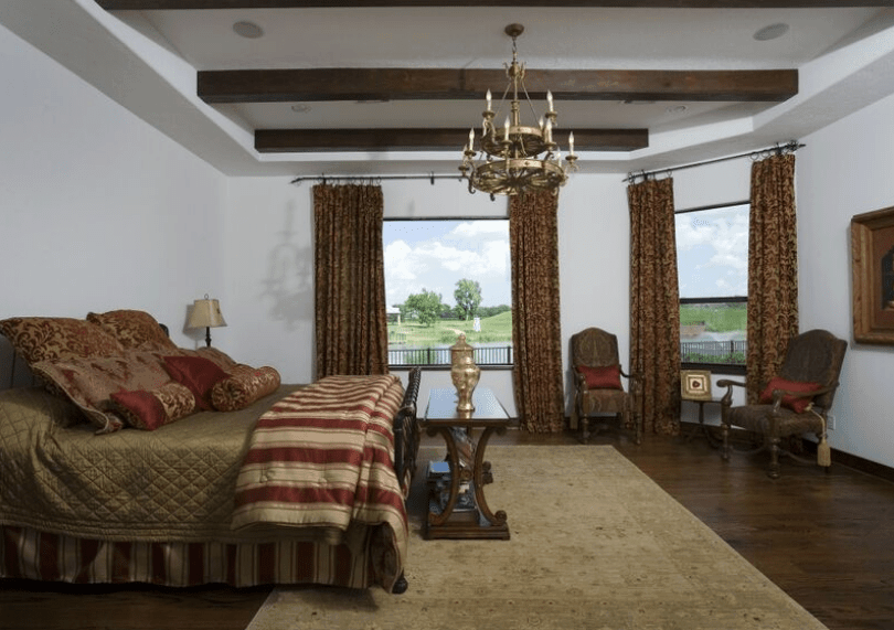 This bedroom is dominated by the chocolate brown hue of the wooden floors, exposed wooden ceiling beams, curtains, table, and French chairs. This is then contradicted by the red stripes of the bedspread, red pillows, and a majestic two-tiered golden chandelier.